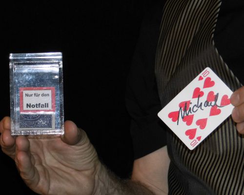 Zauberer Thies hält eine durchsichtige Box in der einen und eine Spielkarte mit dem Namen 'Michael' gekennzeichnet in der anderen Hand